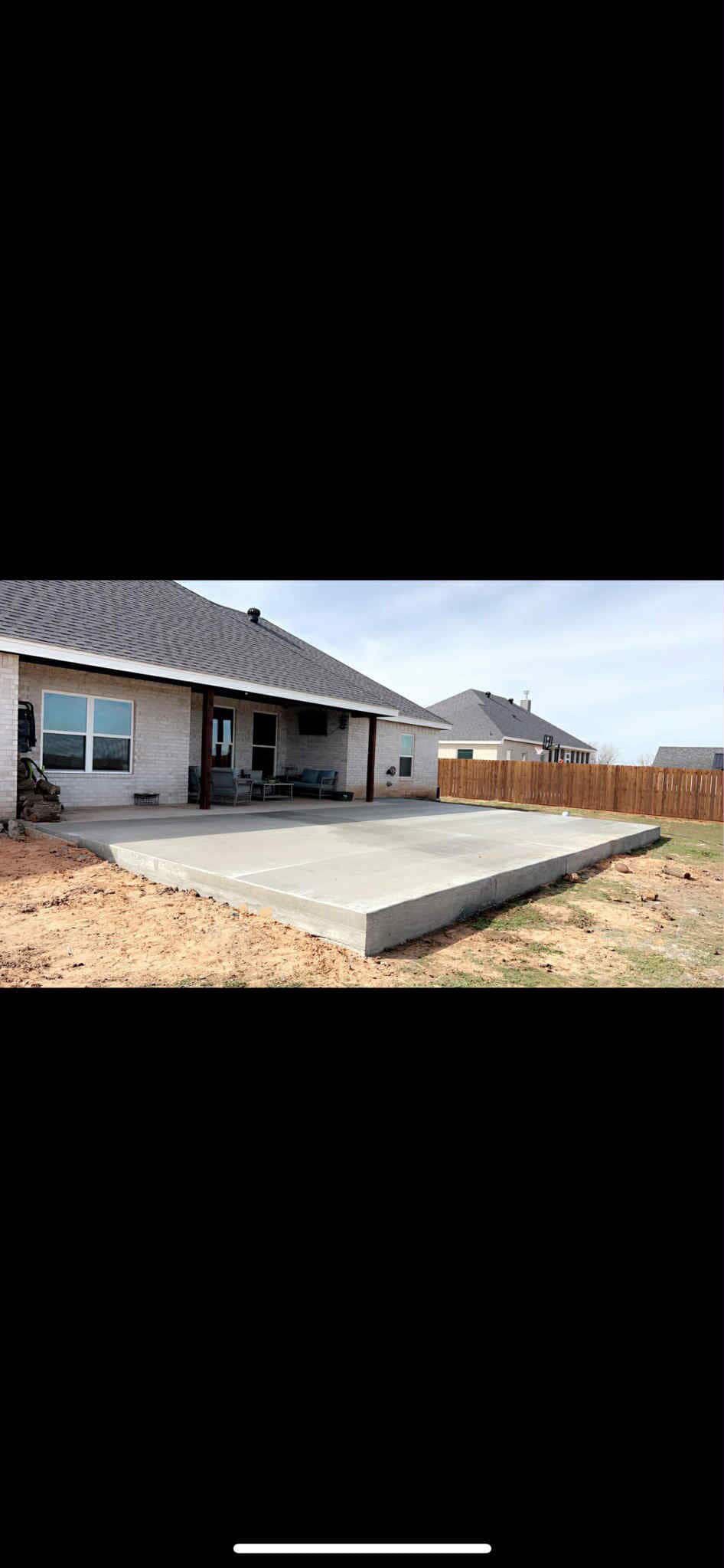A concrete patio is near a home, surrounded by dirt and a wooden fence on a sunny day.