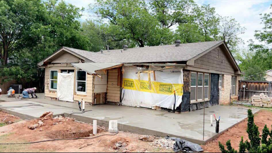 House under construction; concrete patio, exposed framing, siding, and tarp. A worker is seen.