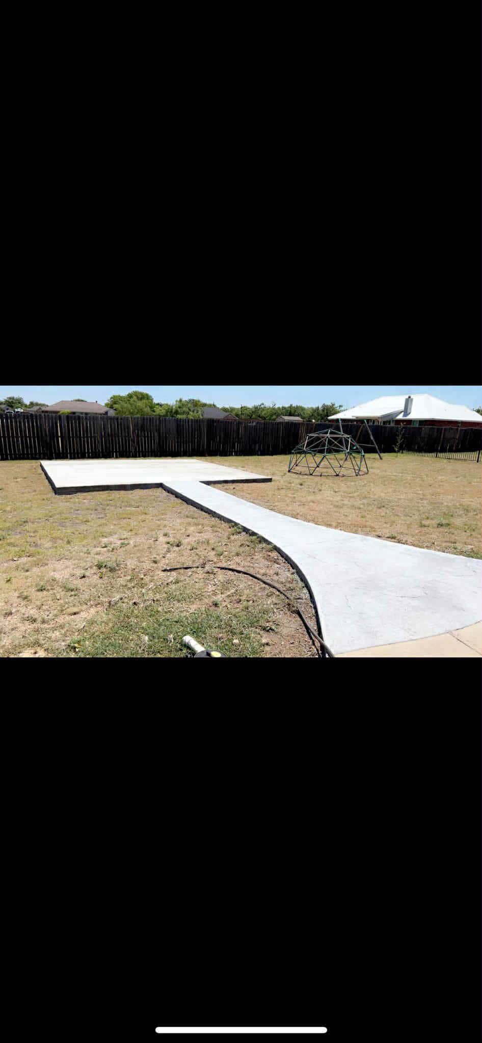 Concrete pathway and patio in a backyard with dry grass and a fence.