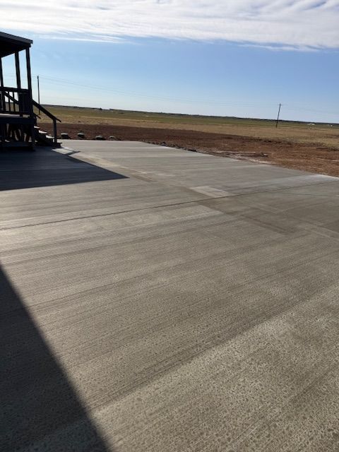 A concrete driveway in a rural setting, leading to a building with wooden steps, under a blue sky.
