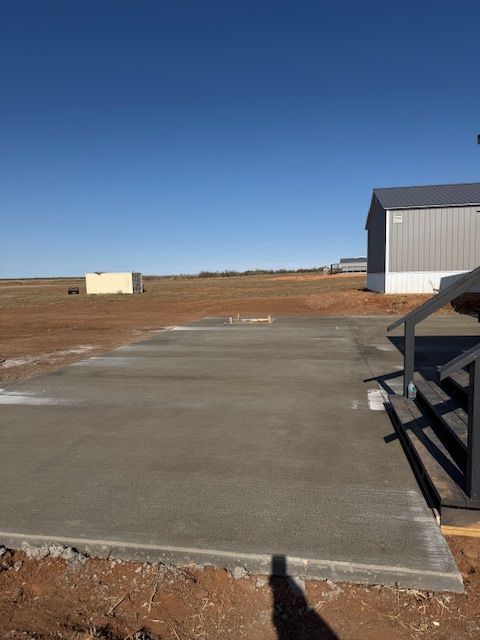 Newly poured concrete slab in an open field, with a metal building in the background under a blue sky.