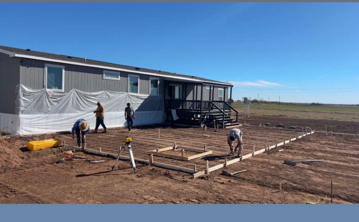 Construction crew building a concrete patio next to a gray mobile home on a sunny day.