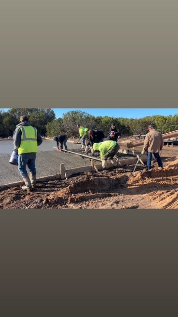 Construction workers pouring concrete outdoors. Some wear safety vests. Brown dirt and green trees visible.