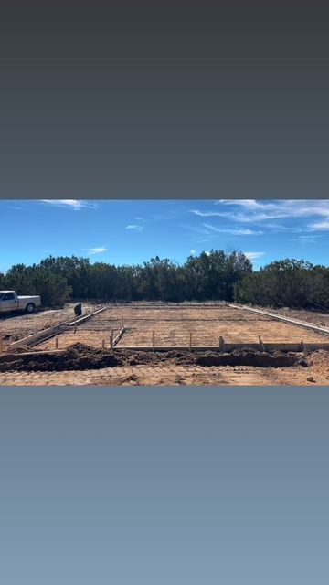 Construction site: foundations outlined on a dirt lot, with trees in the background under a blue sky.