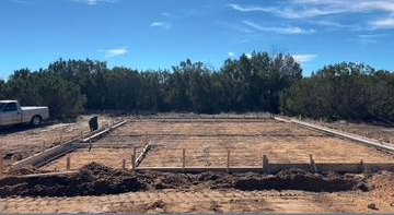 Construction site with wooden forms for a concrete foundation. A truck and trees are visible in the background.