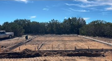 Construction site with concrete forms set on dirt, with trees and blue sky.