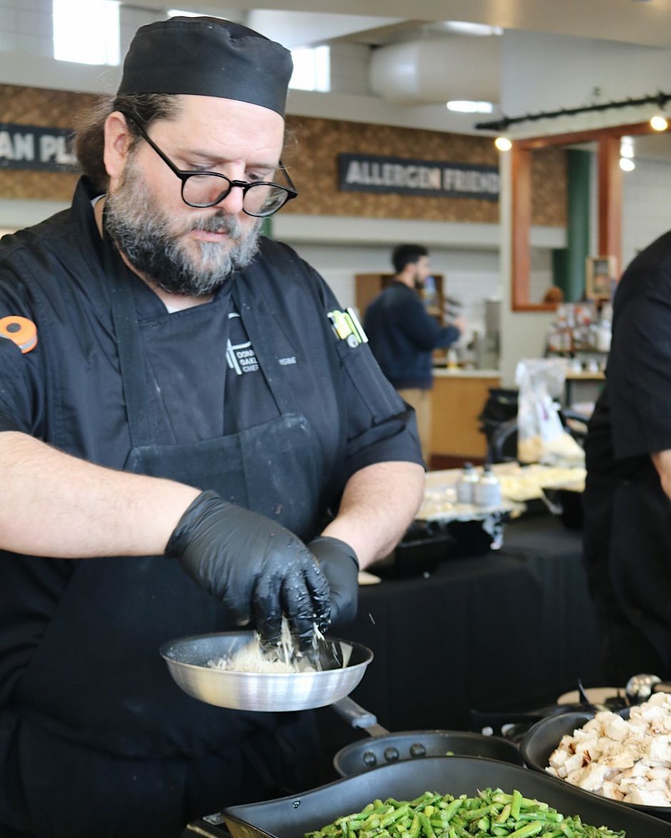 Chef in black uniform cooking, adding ingredient to pan. Indoors, with other food prepared nearby.