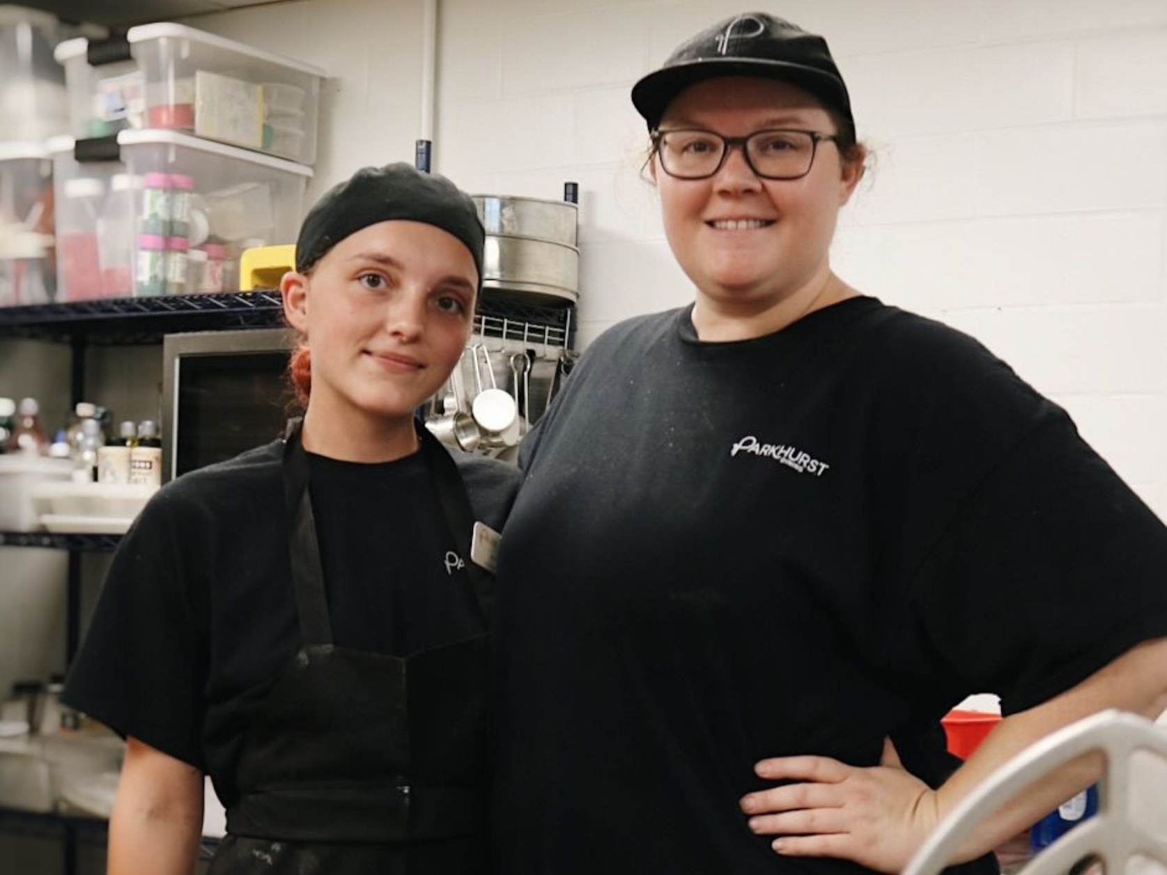 Two chefs in a commercial kitchen with trays of strawberries, smiling, one wearing a hat.