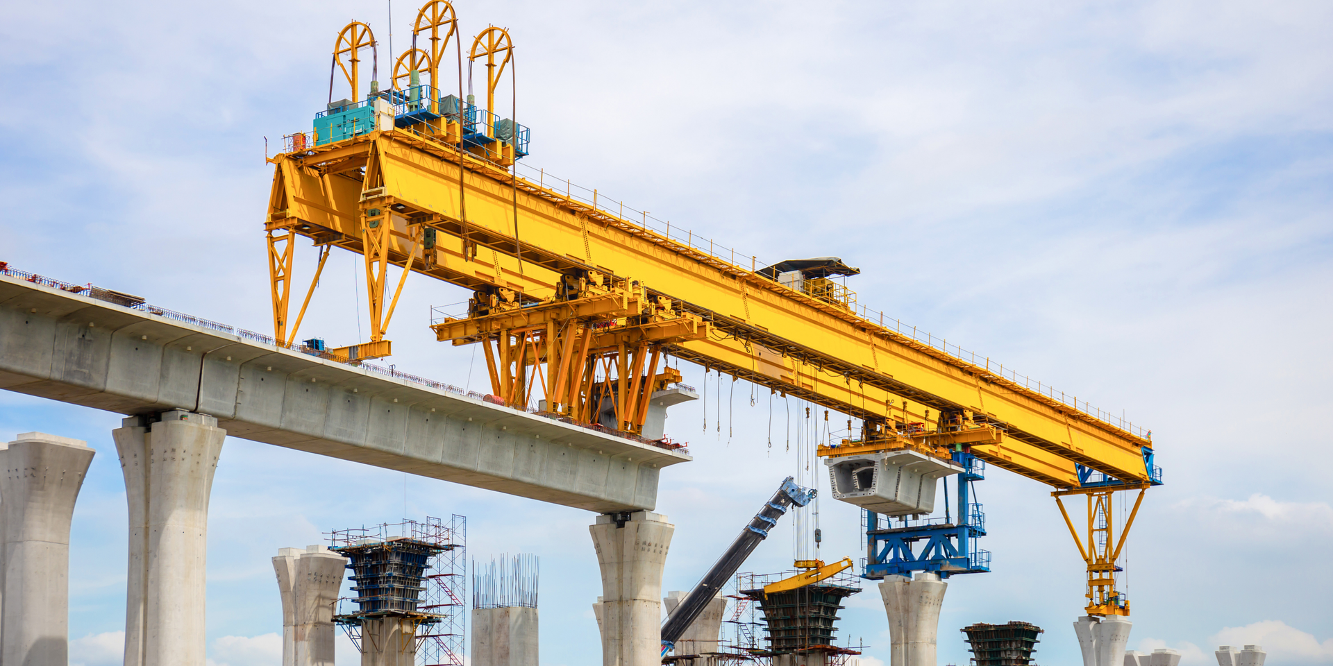 A yellow bridge-launching gantry crane lifts a concrete segment to install it onto support pillars at a construction site.