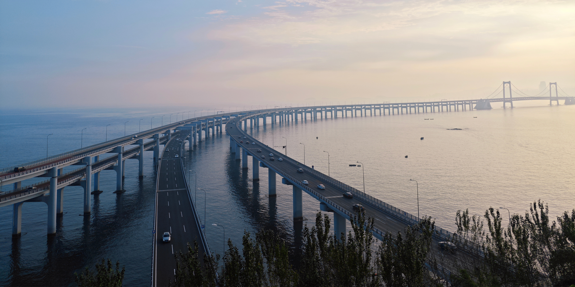 An elevated view of a long, curved bridge spanning calm water during sunset, with traffic moving along the lanes.