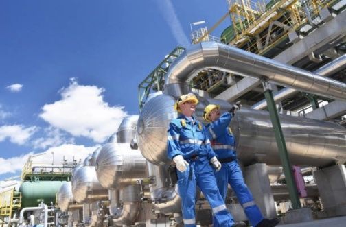 Two men are standing in front of a large pipe in a factory.
