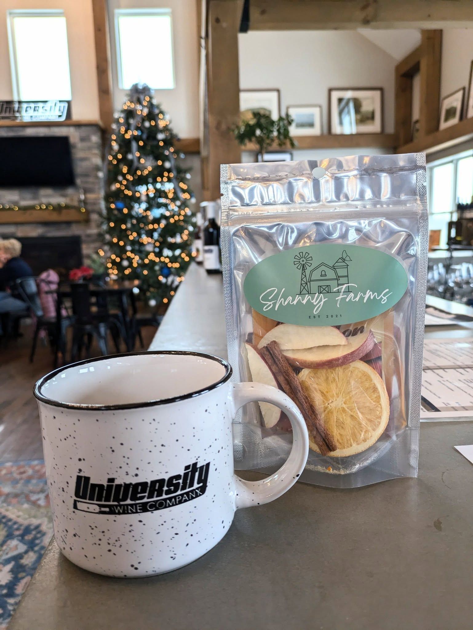 A mug and a bag of fruit chips are on a table in front of a christmas tree.