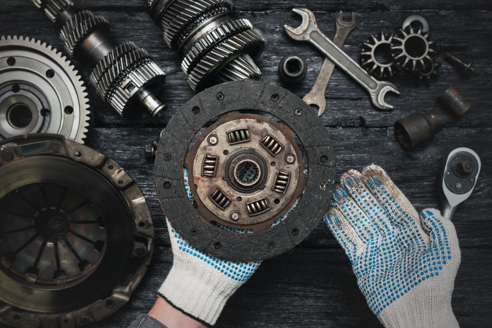 Hands holding a car clutch disk surrounded by auto repair tools and parts on a dark workbench | Quality Auto Repair Tire & Auto Sales