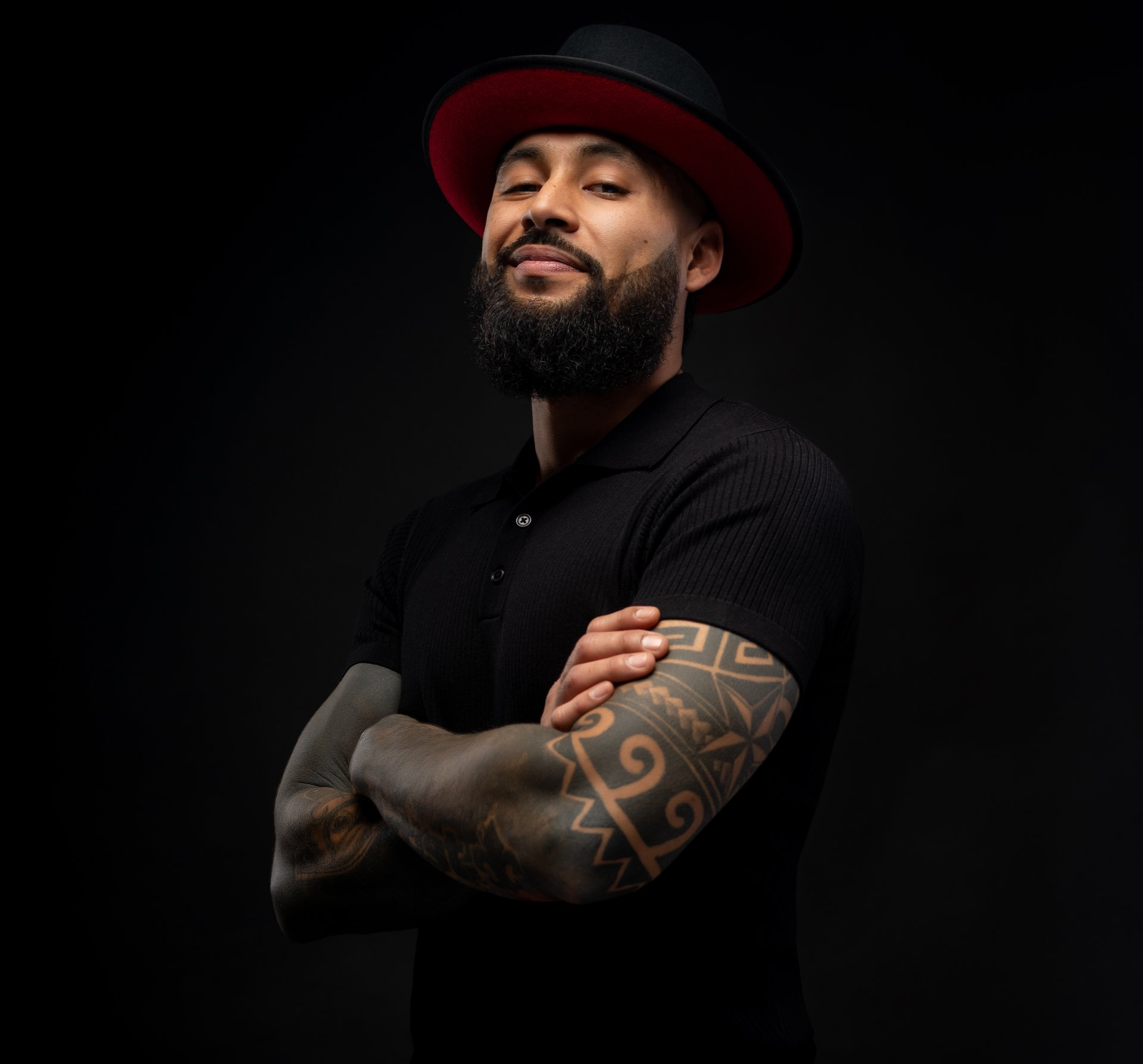 A man with a beard and tattoos on his arms is wearing a hat and a black shirt.