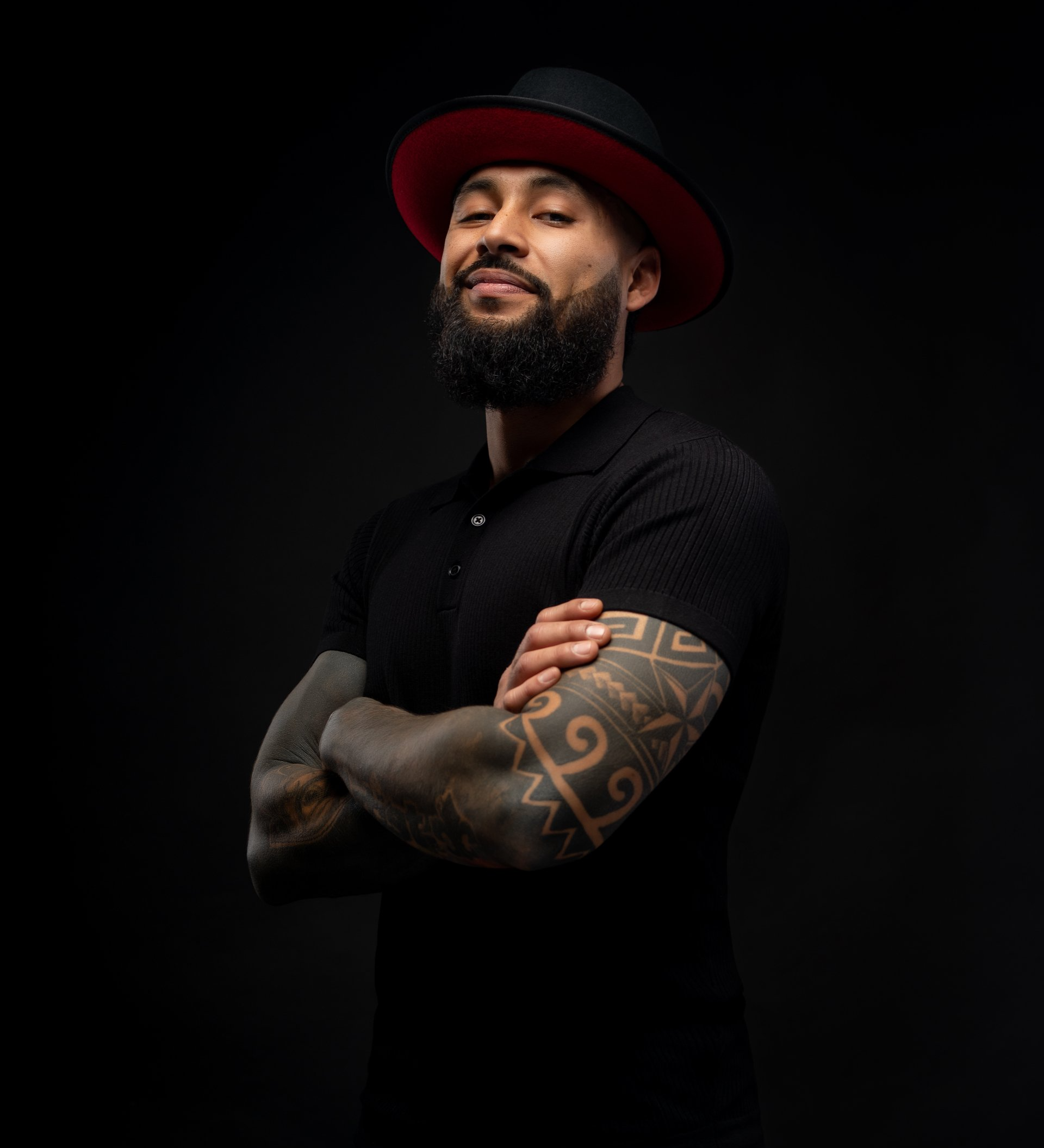 A man with a beard and tattoos is wearing a hat and a black shirt.