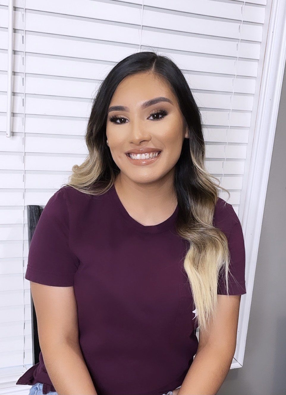 A woman in a purple shirt is smiling while sitting in front of a window.