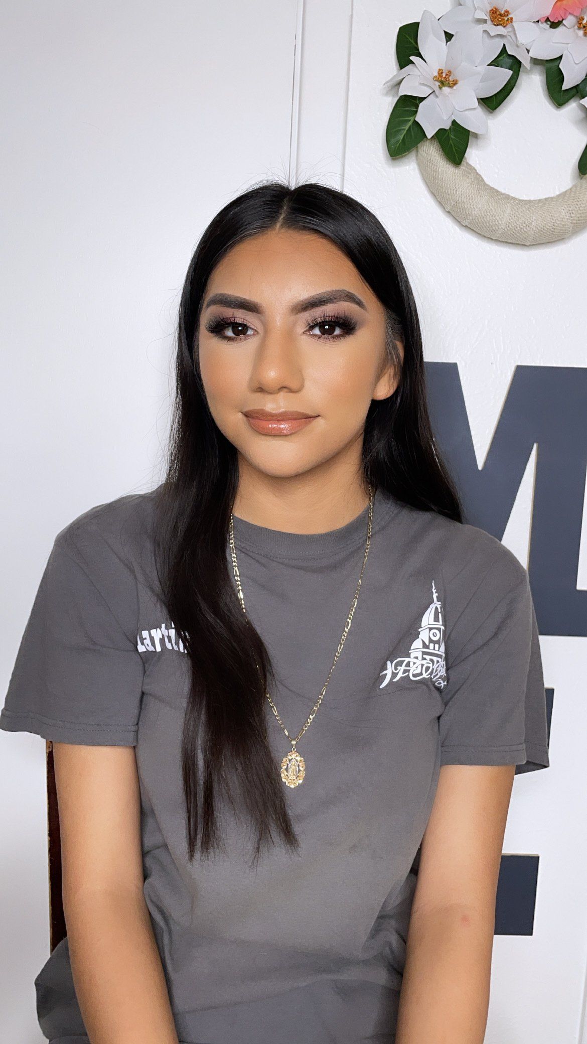 A woman wearing a gray t-shirt and a necklace is sitting in front of a wall with flowers.