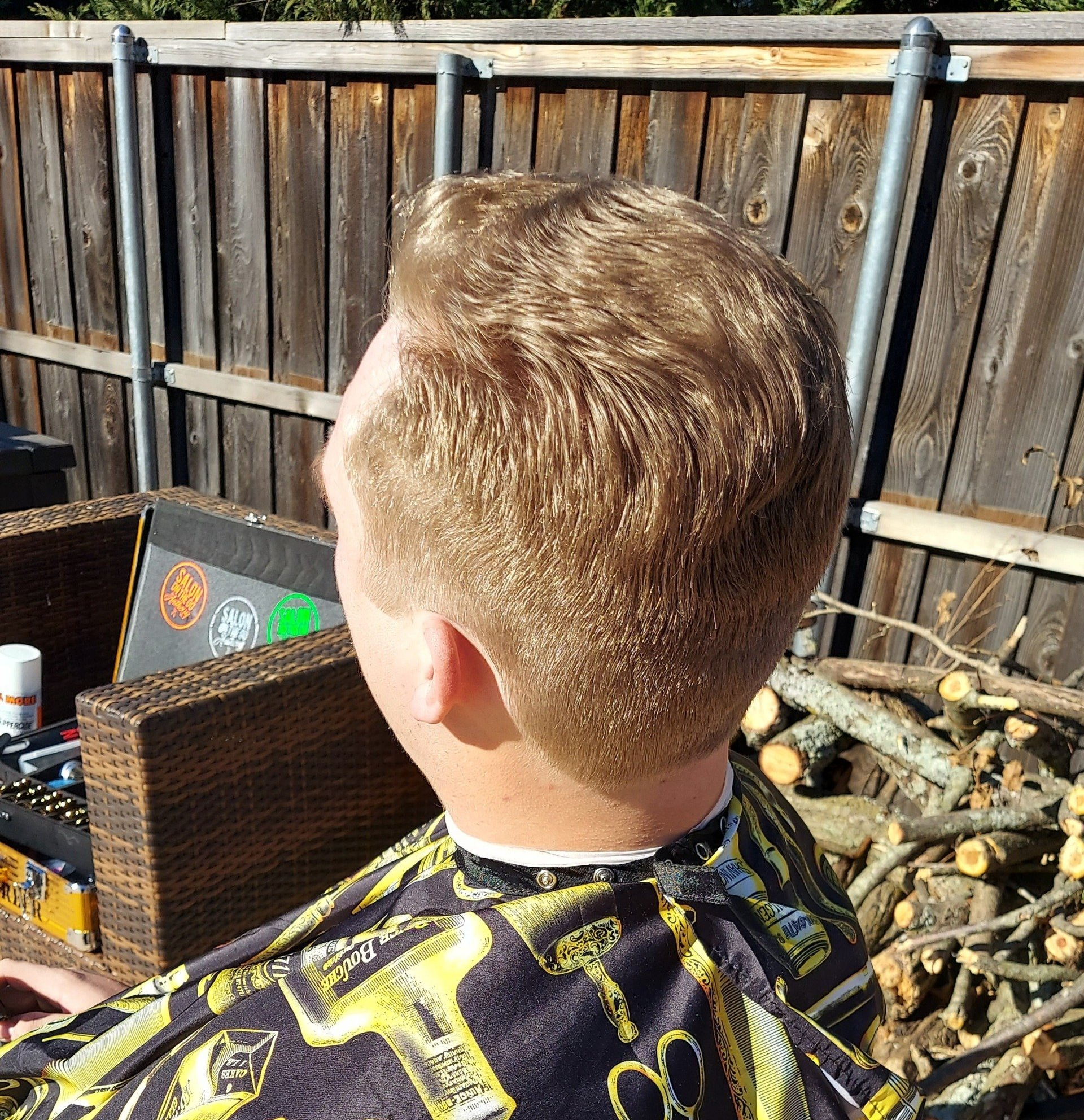 A man is getting his hair cut by a barber in front of a wooden fence.