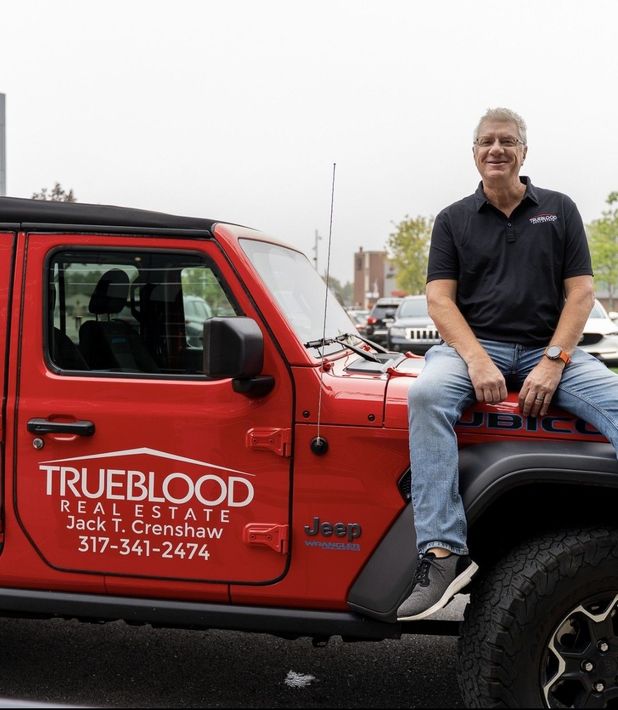 Man in jeans and black shirt sits on a red Jeep; Trueblood Real Estate logo on door.