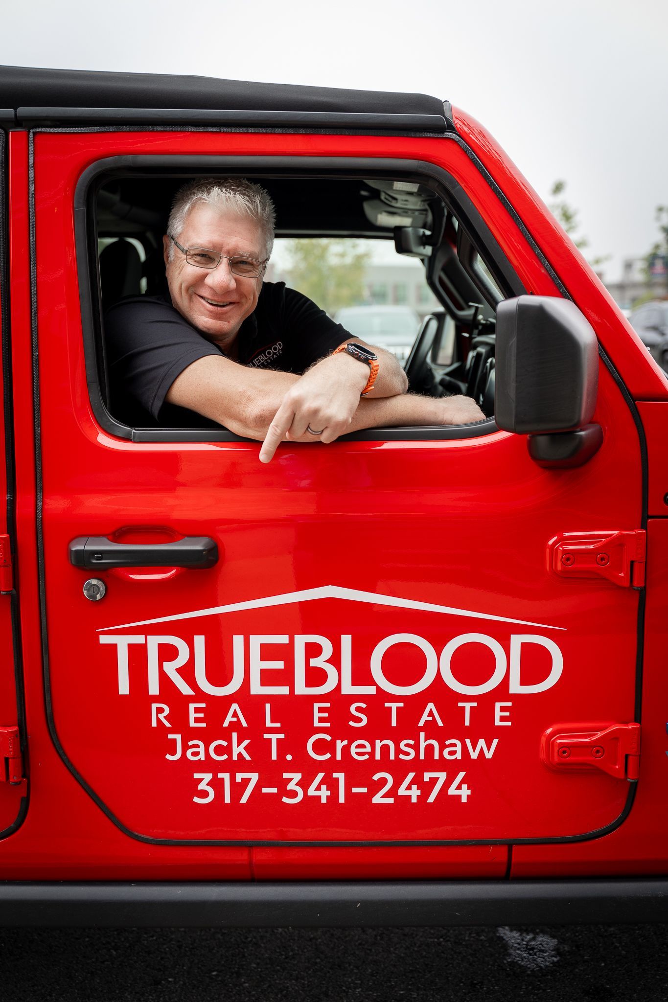 Man in jeans and black shirt sits on a red Jeep; Trueblood Real Estate logo on door.