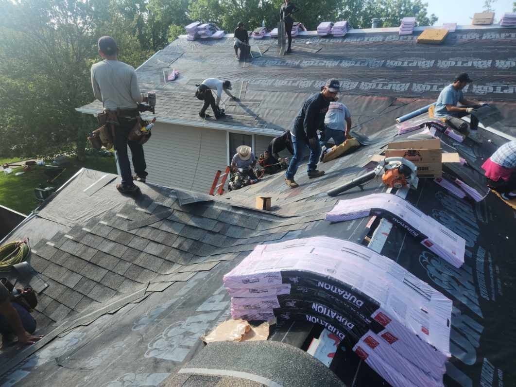 Roofers installing shingles on a residential roof, stacks of materials visible.