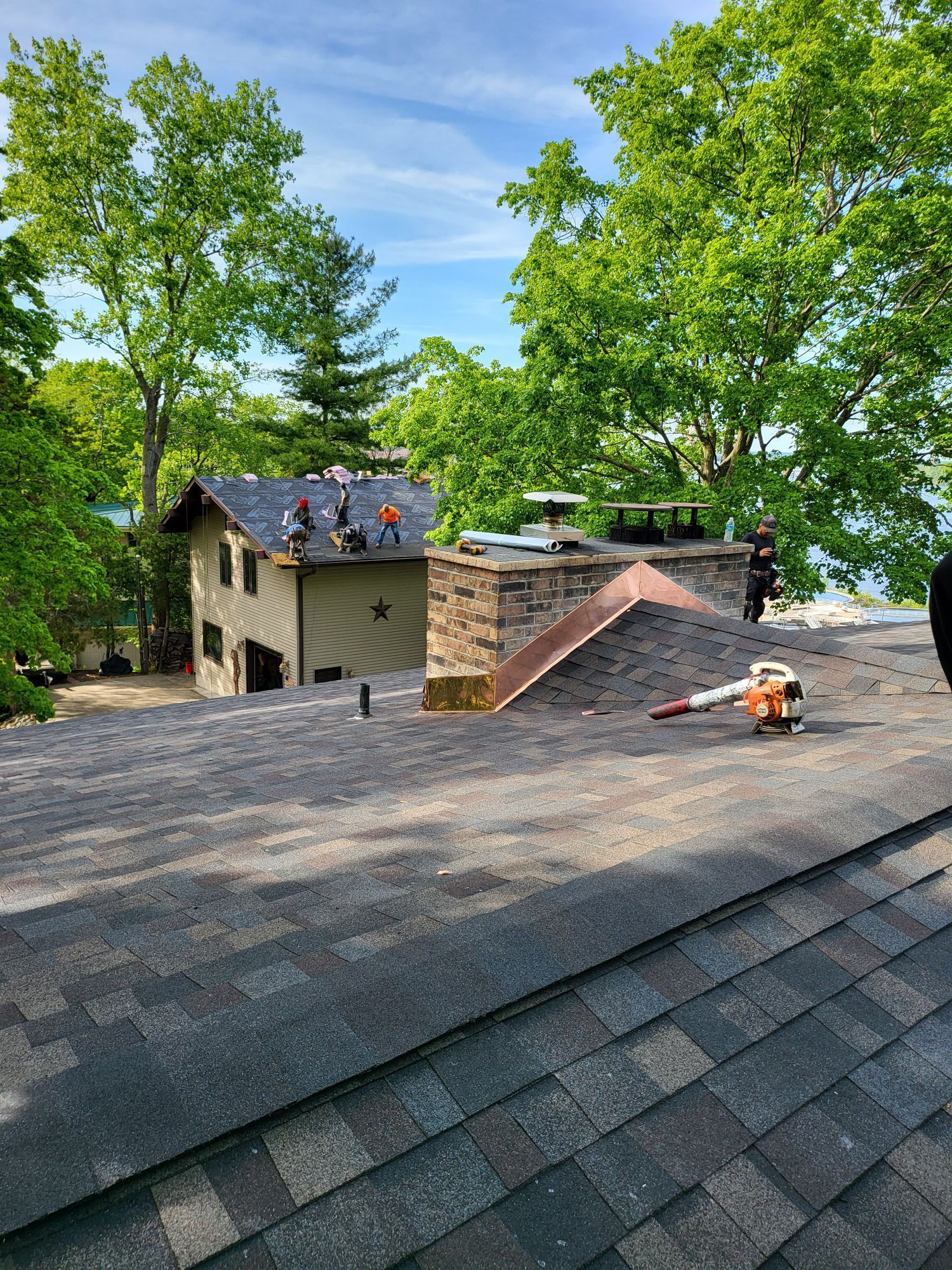 Workers on a roof with a chimney, blowing debris; another house in the background near trees and water.