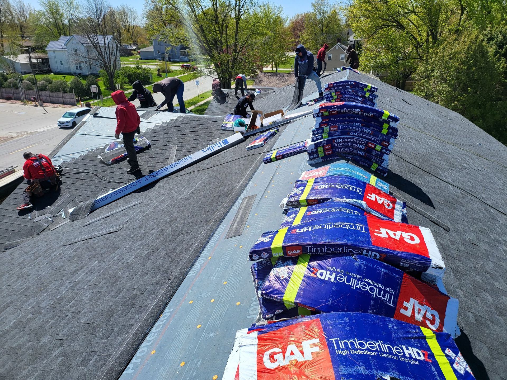 Roofers installing shingles on a house roof. Blue and red shingle bundles are present along the roofline.