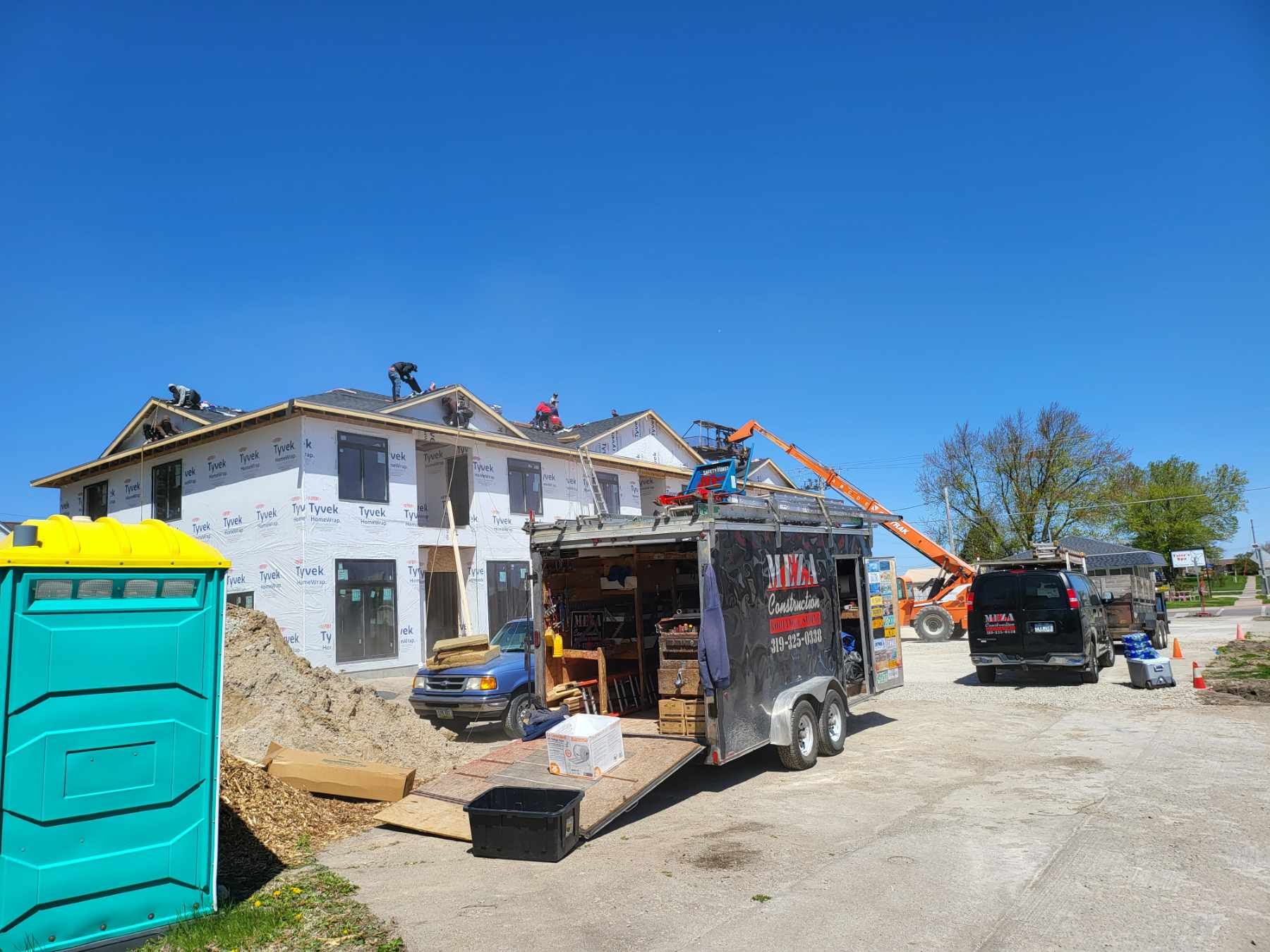 Construction site with workers on a roof, tools in a trailer, portable toilet, and equipment under a clear blue sky.