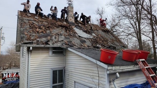 People removing shingles from a house roof; red buckets and ladder visible. Overcast day.