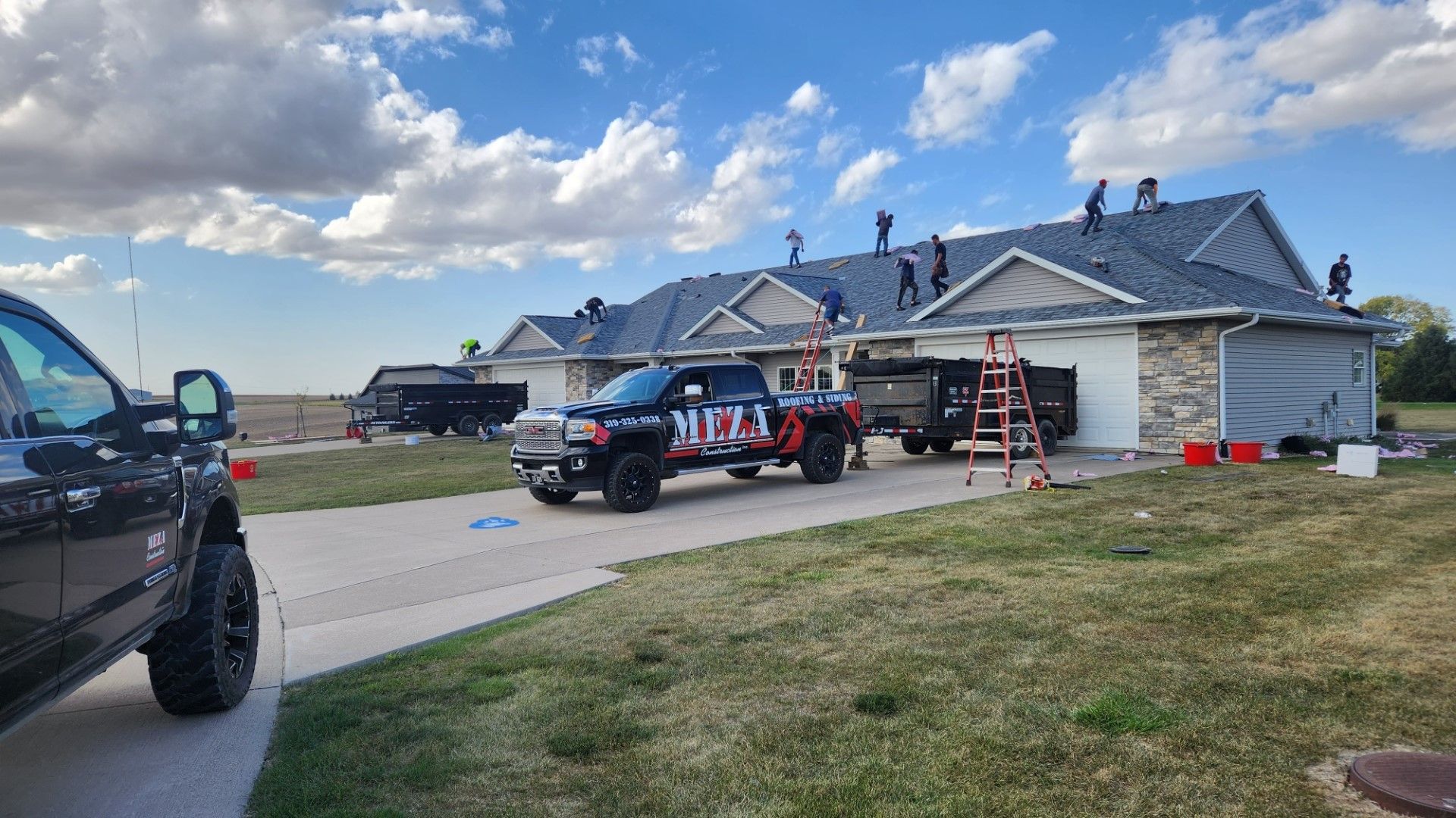 Roofers on a residential roof, with trucks and dumpsters in the driveway on a sunny day.