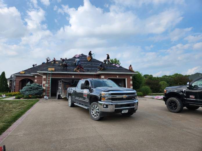 Roofers working on a house roof. A truck and trailer are parked in the driveway. Blue sky with clouds.