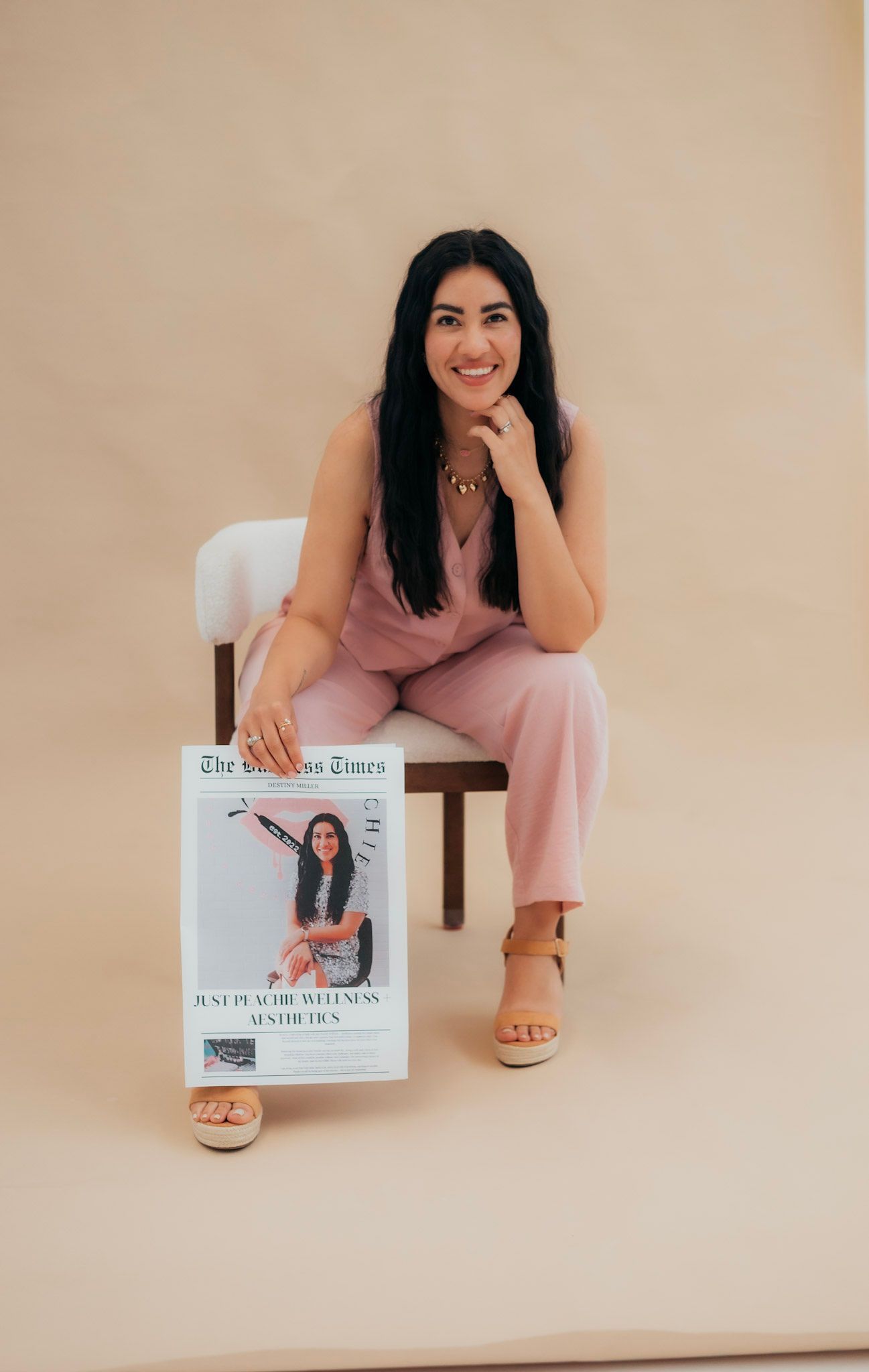 Woman in pink outfit smiles while holding a magazine. Sitting on a chair in front of a tan backdrop.