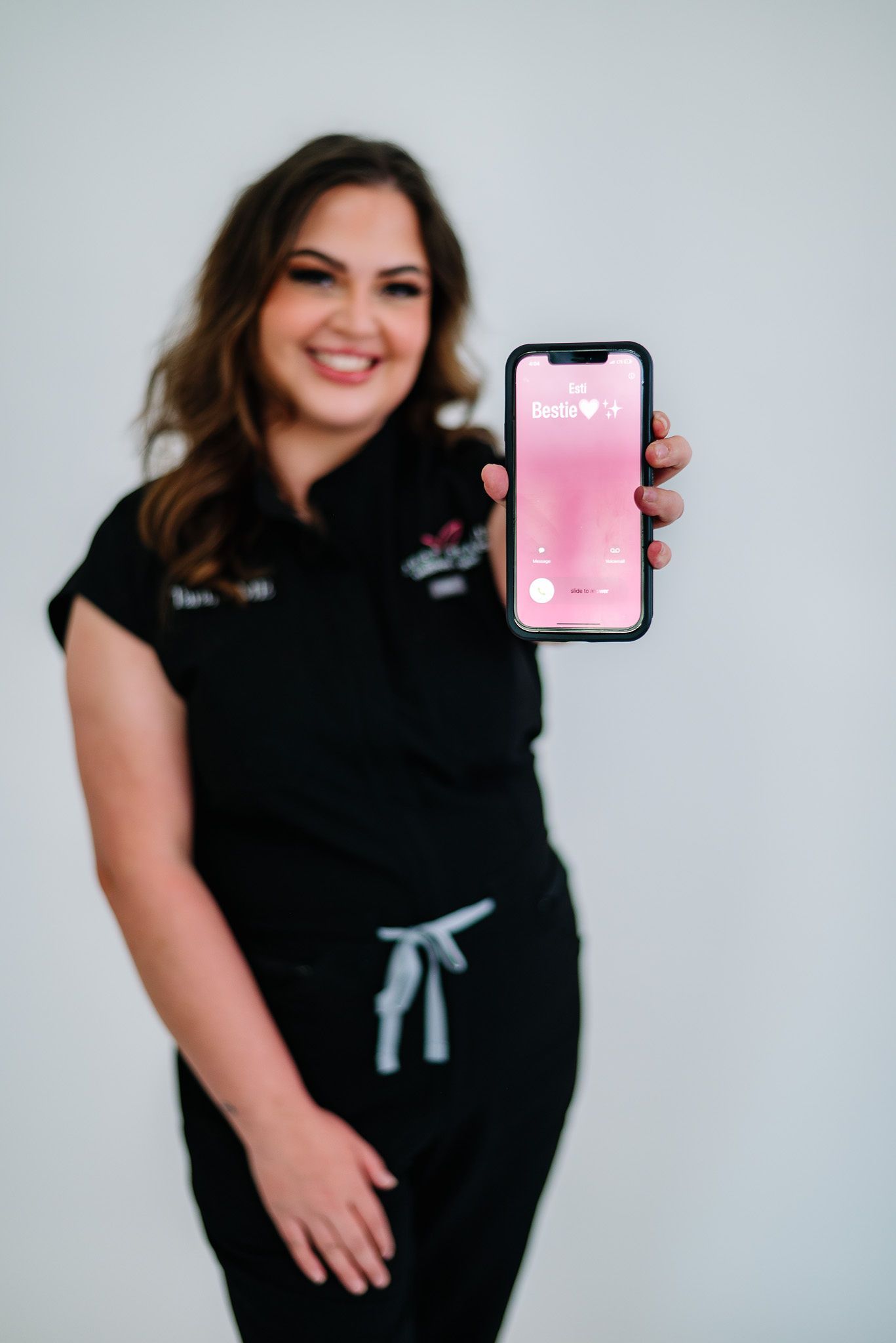 Woman in black scrubs smiles, holding up a phone with a pink screen against a white background.