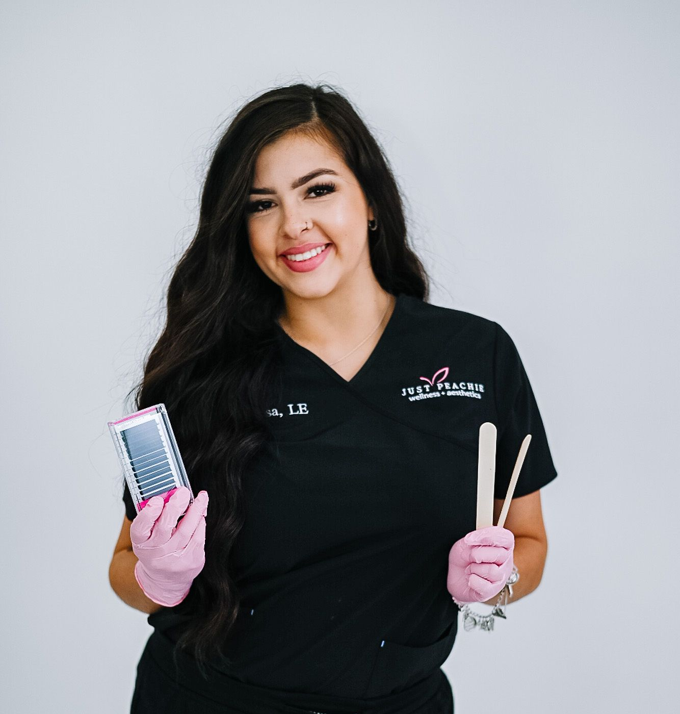 Woman in black scrubs holds waxing tools and smiles at the camera.