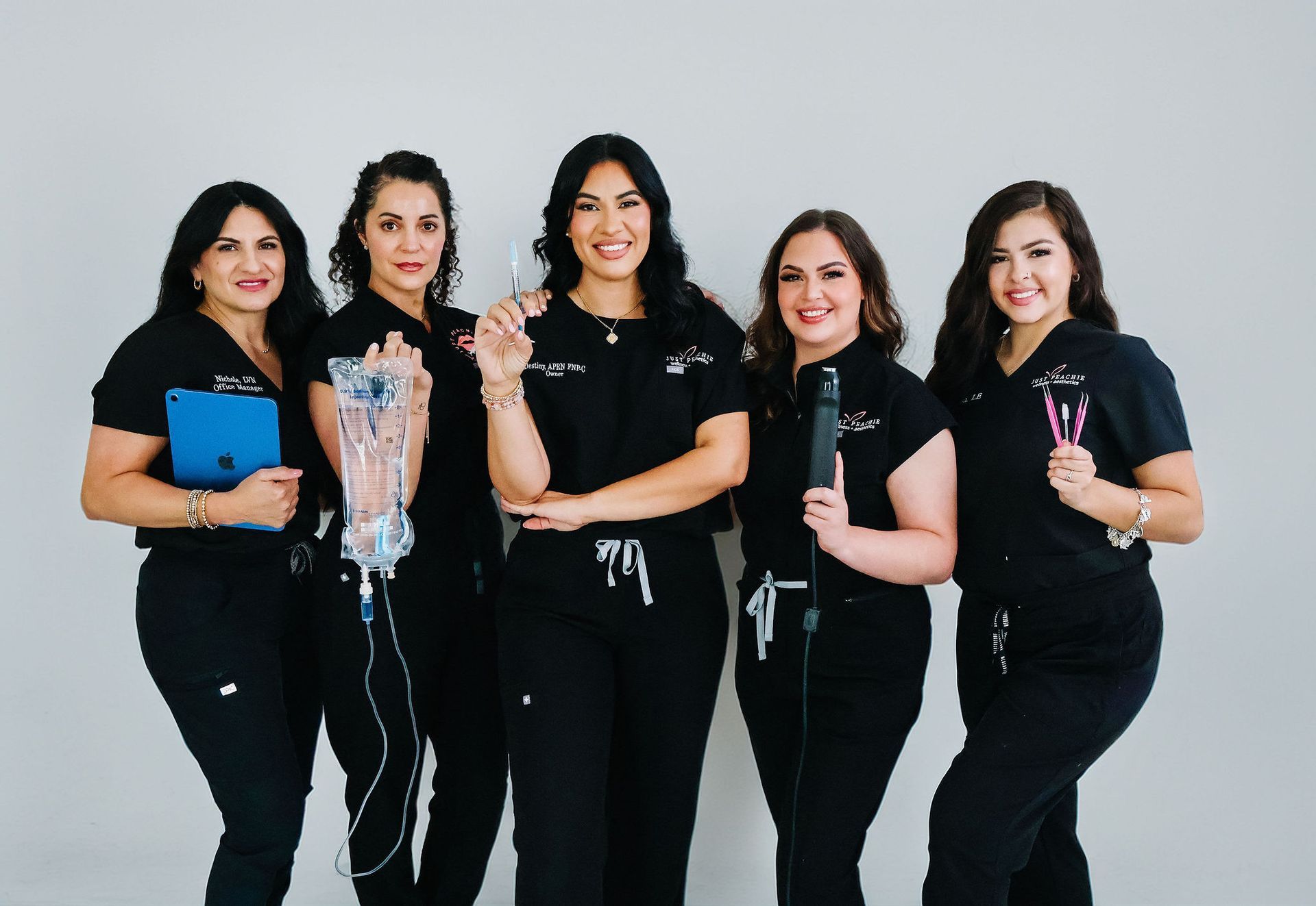 Five women in black scrubs, smiling and holding medical tools.