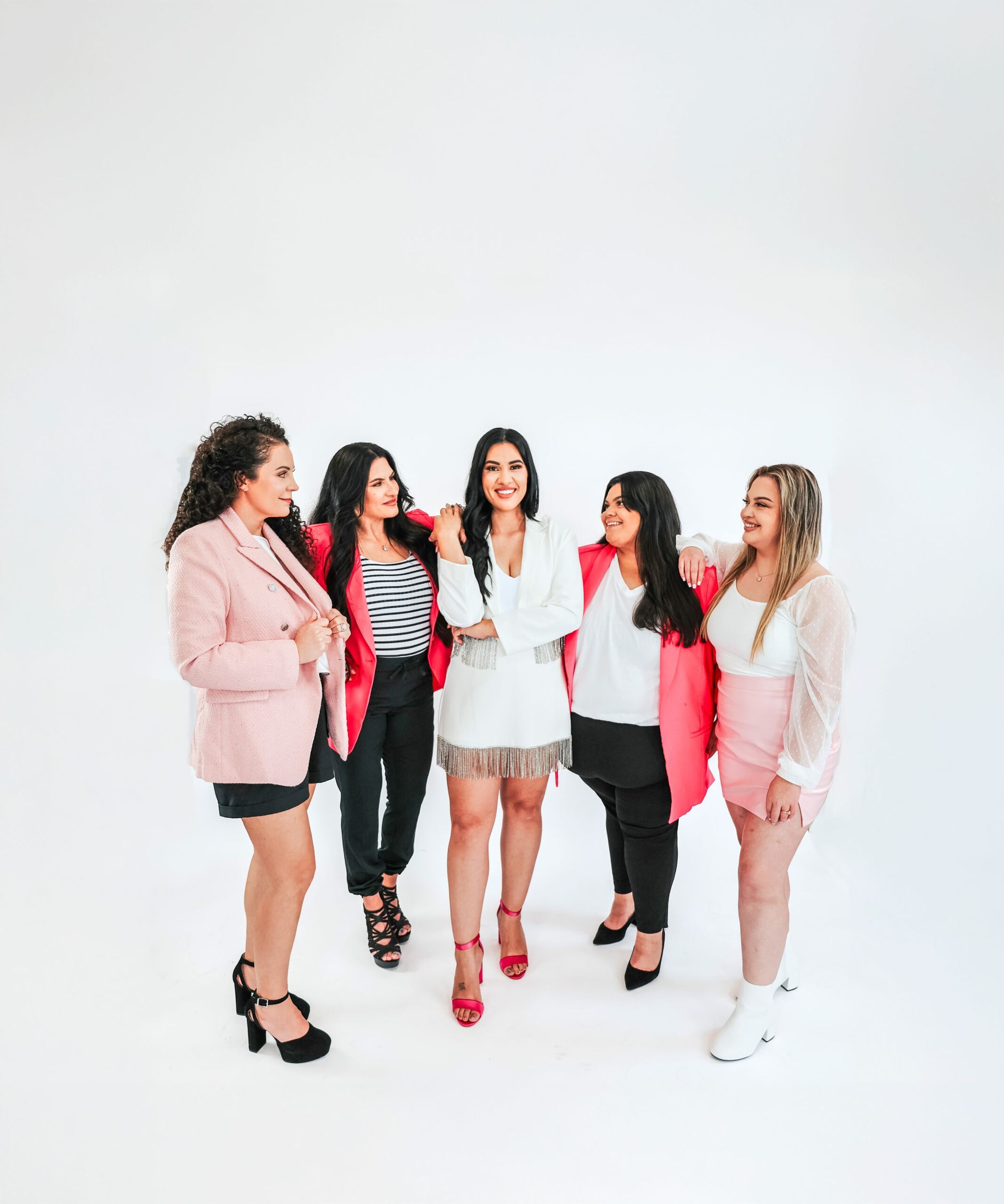 Five women in bright outfits posing and smiling against a white background.