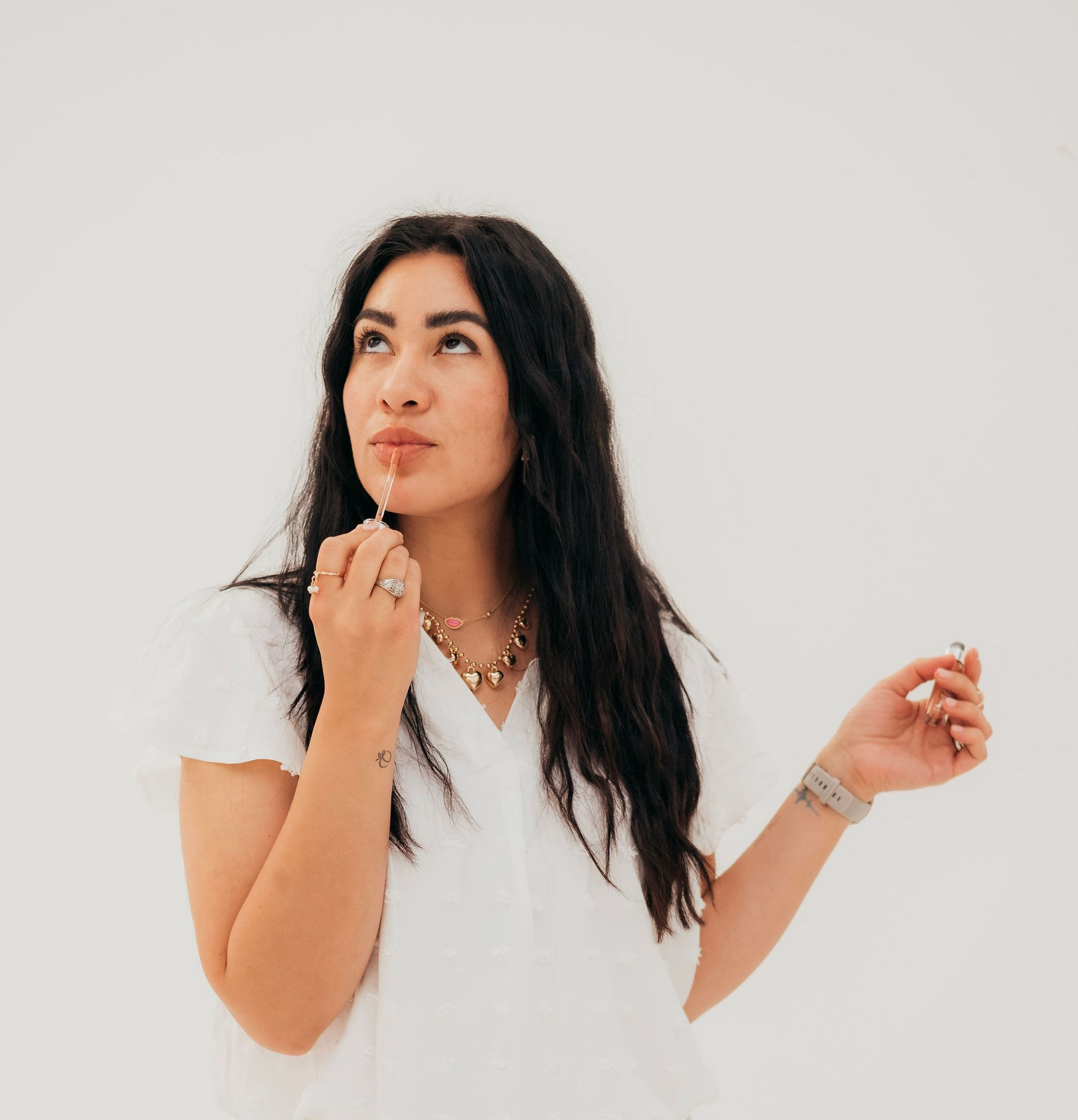 Woman with long dark hair looking up, applying lip gloss, wearing white top and jewelry, white background.