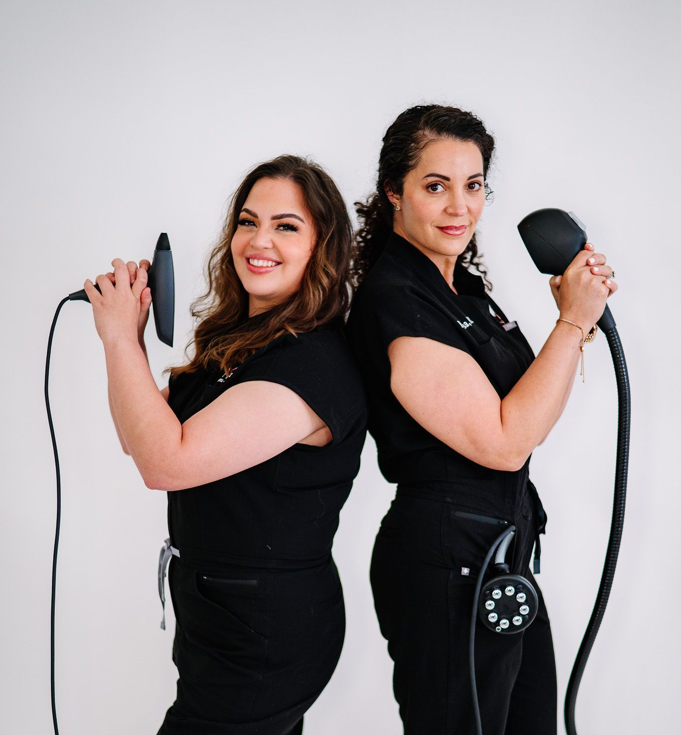 Two women in black scrubs holding body contouring devices, smiling, standing back-to-back in front of a white background.