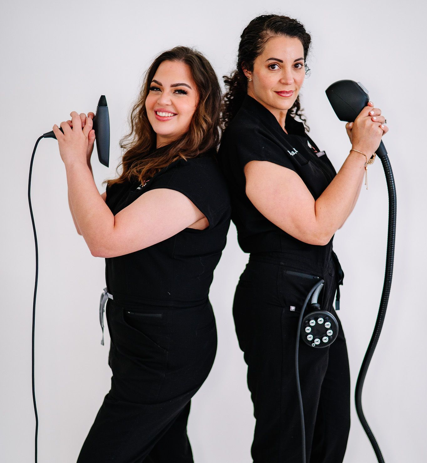 Two women in black scrubs, holding medical devices, smiling, standing back-to-back against a white background.