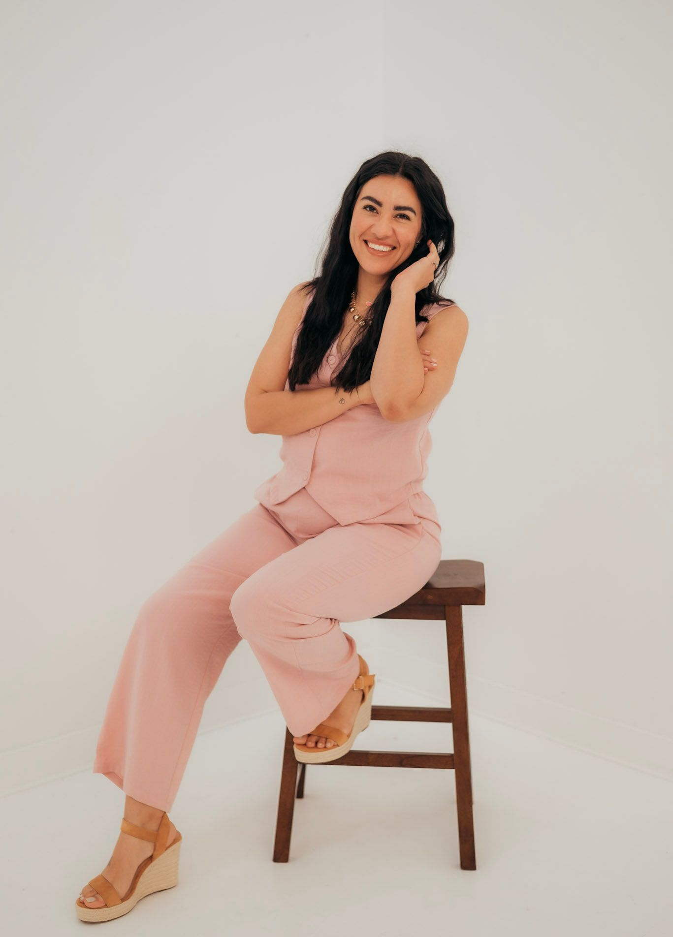 Woman in pink jumpsuit smiling, seated on a wooden stool, hand in hair. White background.