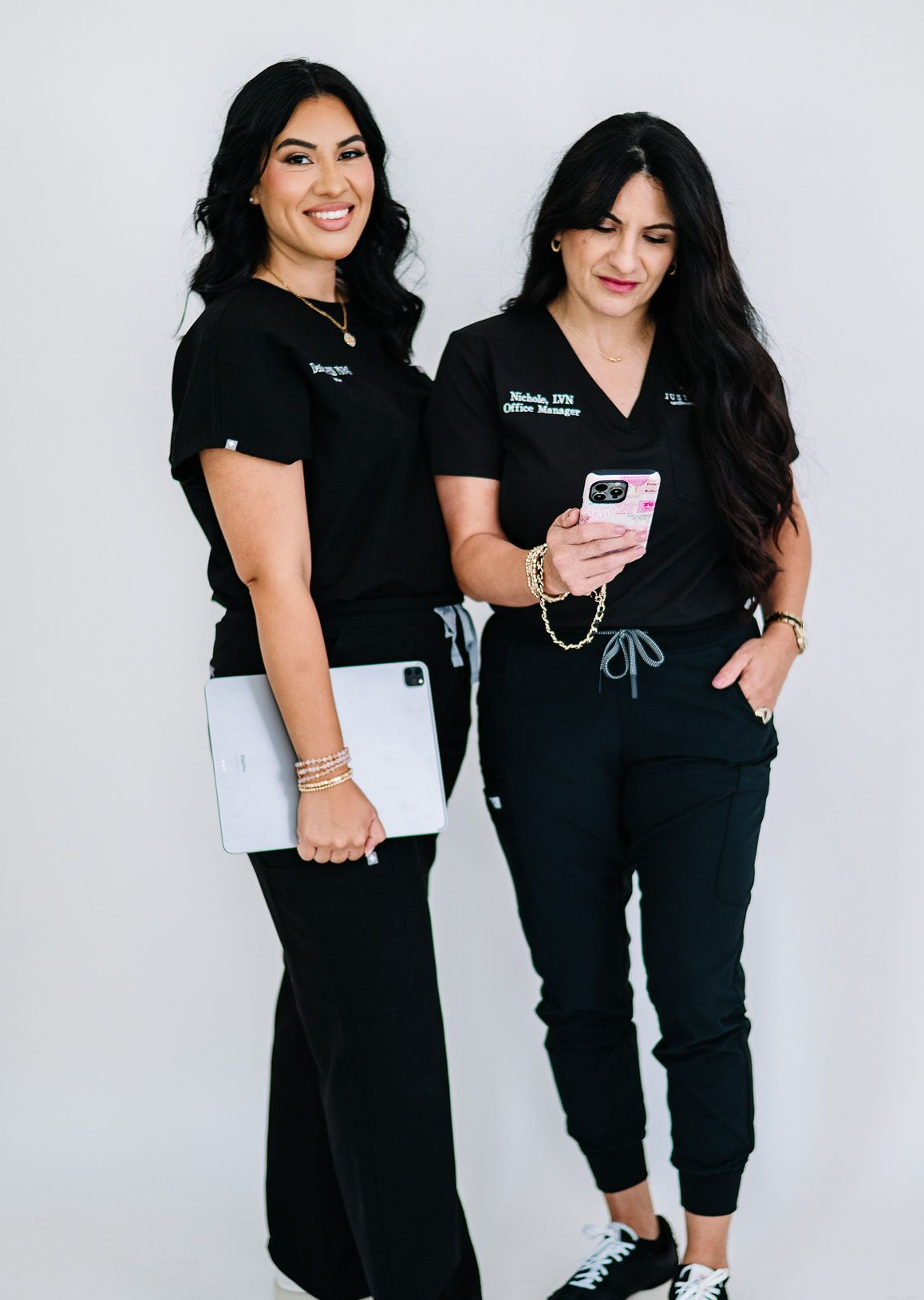 Two women in black scrubs, one holding a laptop, the other with a phone, smiling.