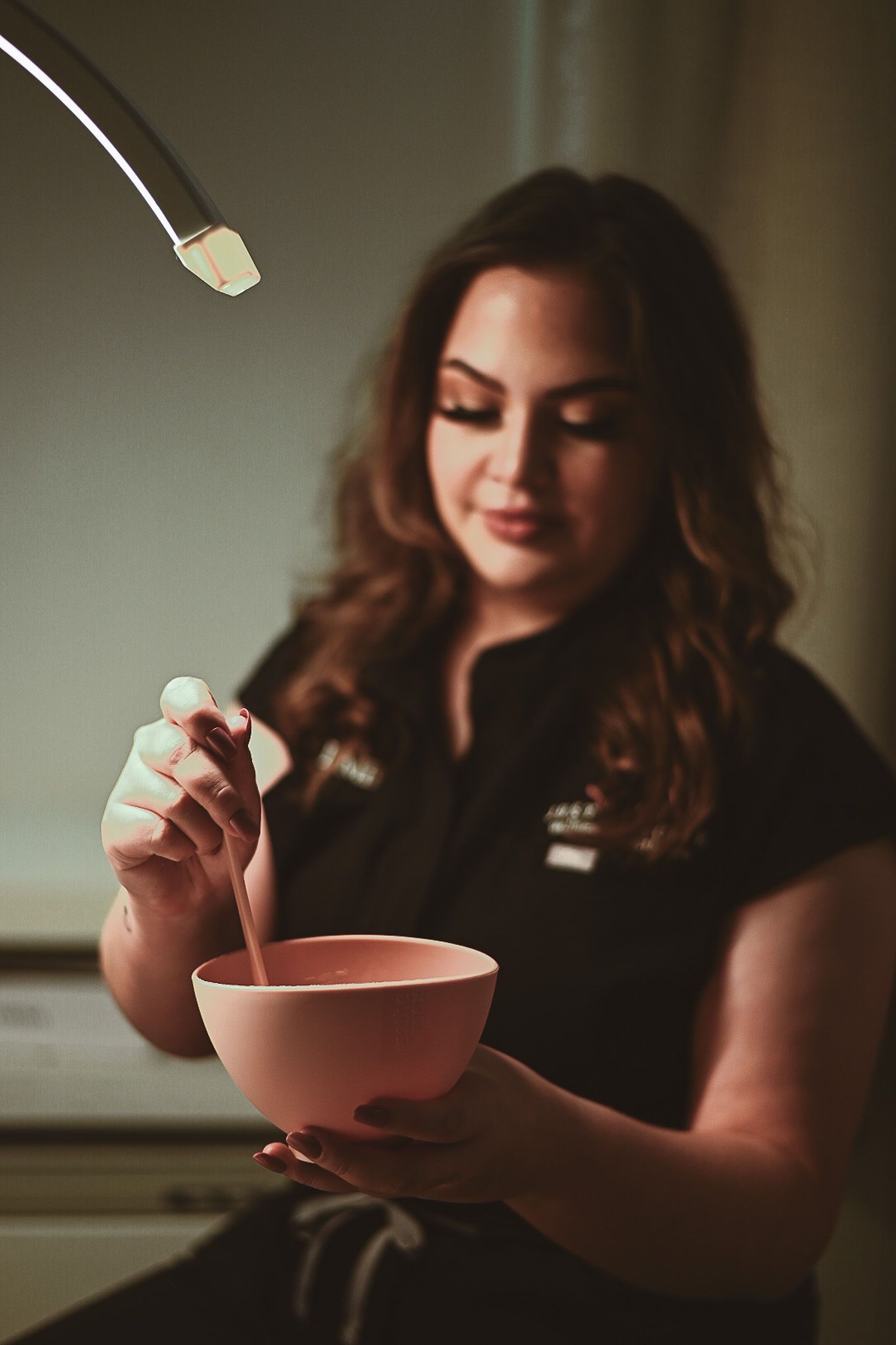 Woman in black shirt stirring pink substance in a bowl under a lamp.
