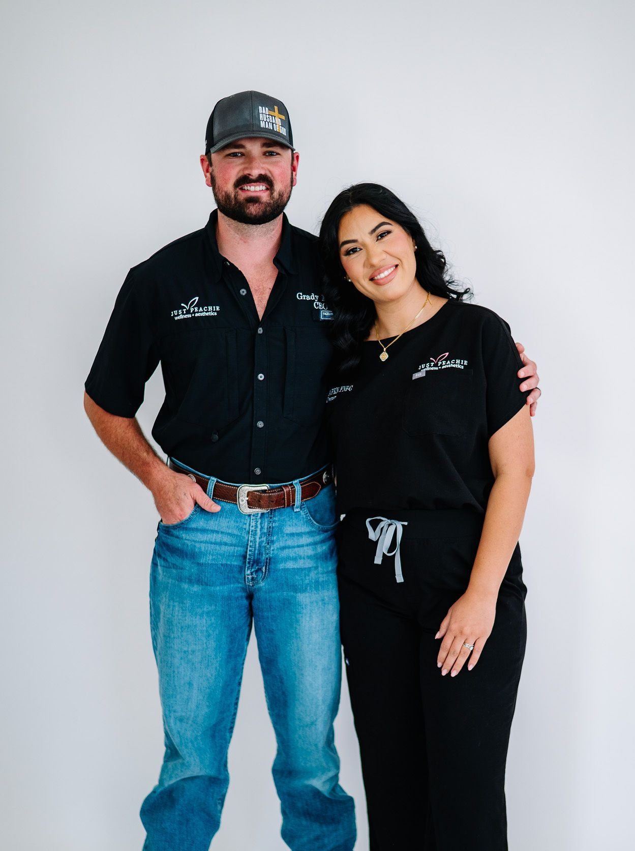 Man and woman standing, smiling. Man in jeans, woman in scrubs. White backdrop.