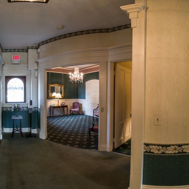 Hallway with patterned wallpaper, leading to a room with a chandelier and furniture.
