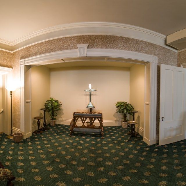 Interior of a chapel with a cross, plants, and green patterned carpet. A door and a lamp are on either side.