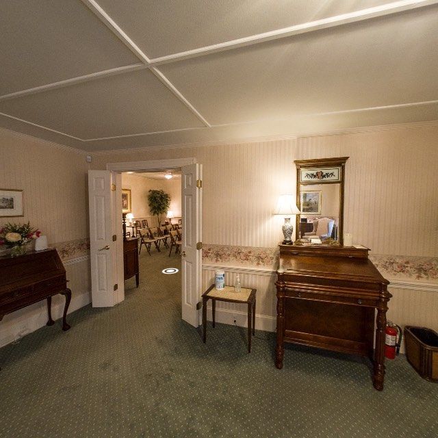 A room with green carpet, wooden furniture, and a doorway leading to another room; likely a funeral home.