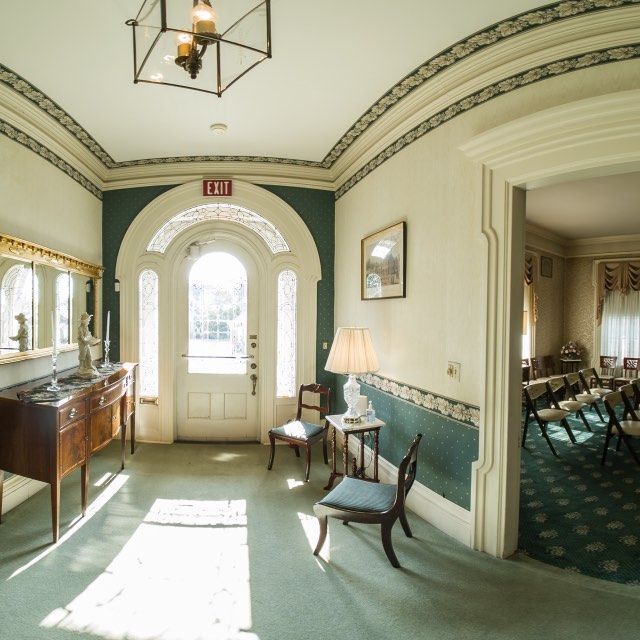 Hallway with ornate trim, green walls, antique furniture, and chairs, and a doorway to a room with chairs.