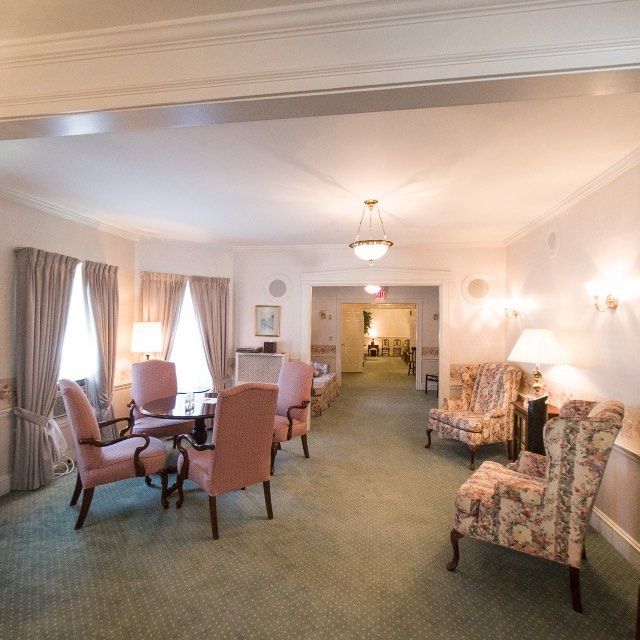 A waiting room with pink chairs around a table and floral armchairs. Soft lighting and green carpet.