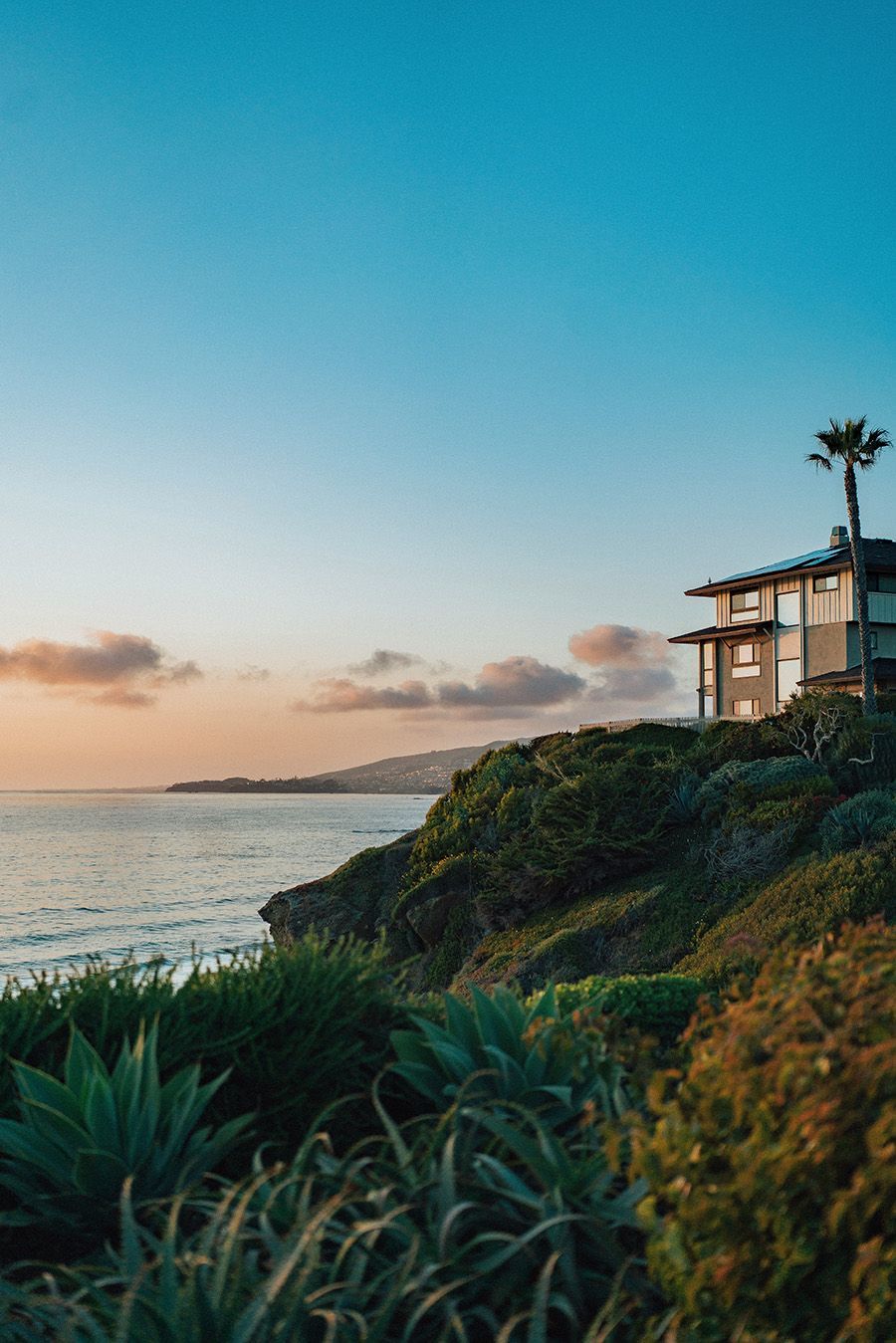 Oceanfront house on a cliff at sunset; blue sky, green foliage, calm water.