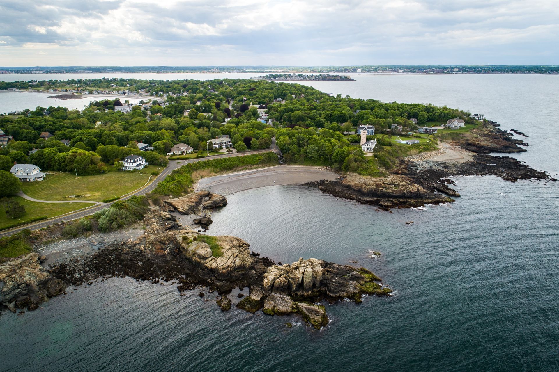 Coastal view of rocky shoreline, trees, houses, and water under a cloudy sky.